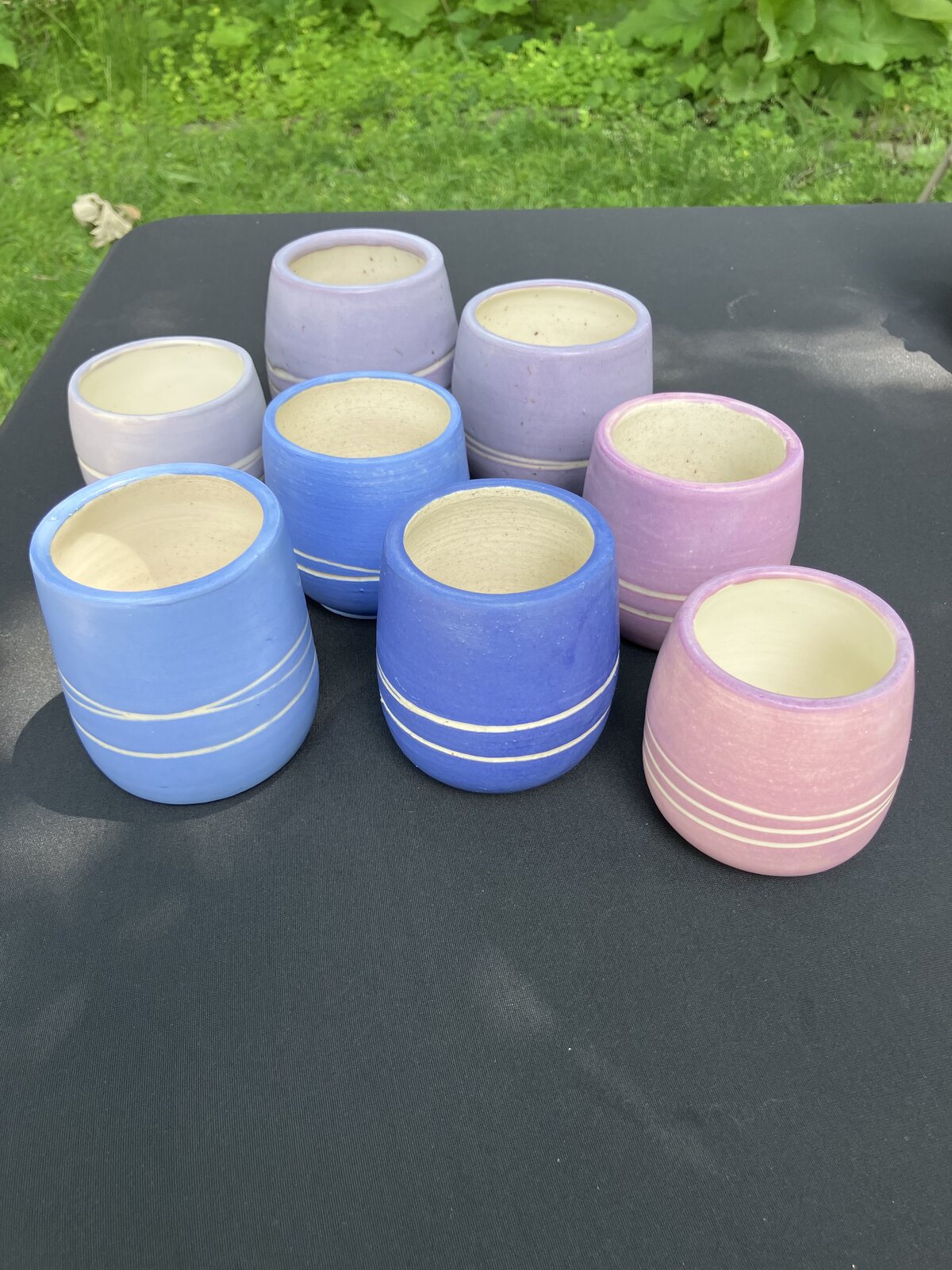 A row of handmade ceramic cups in lavender, blue, and pink glazes on an outdoor table